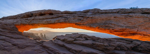 Panoramic fine art photograph of Mesa Arch at sunrise, Canyonlands National Park, Utah — the sandstone arch glowing deep amber and orange at first light.
