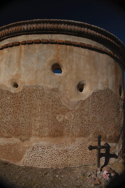 Fine art photography by Jess R. Lee of an adobe mission wall with circular openings and a wooden cross in the foreground.