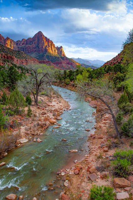 The Virgin River curves through a rocky valley toward a glowing red mountain peak under a dramatic, cloudy sky.