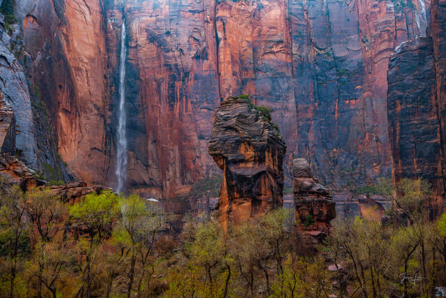 A tall, thin waterfall drops down a massive red sandstone cliff behind dark, weathered rock spires and green desert vegetation.