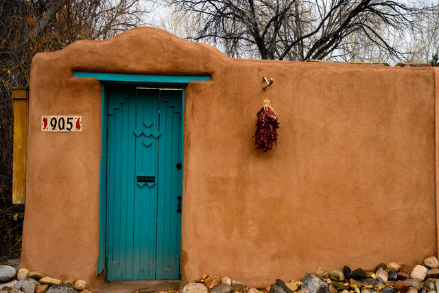 Fine art landscape photography of a vibrant turquoise door with red chile ristras hanging on a textured adobe wall in Santa Fe, New Mexico, by Jess Lee.