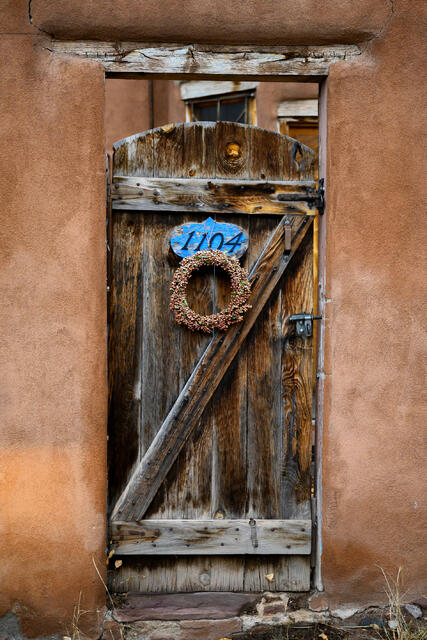 Fine art photography print of a rustic, weathered wood door set in a textured adobe wall in Santa Fe, New Mexico, featuring Southwest architectural details.