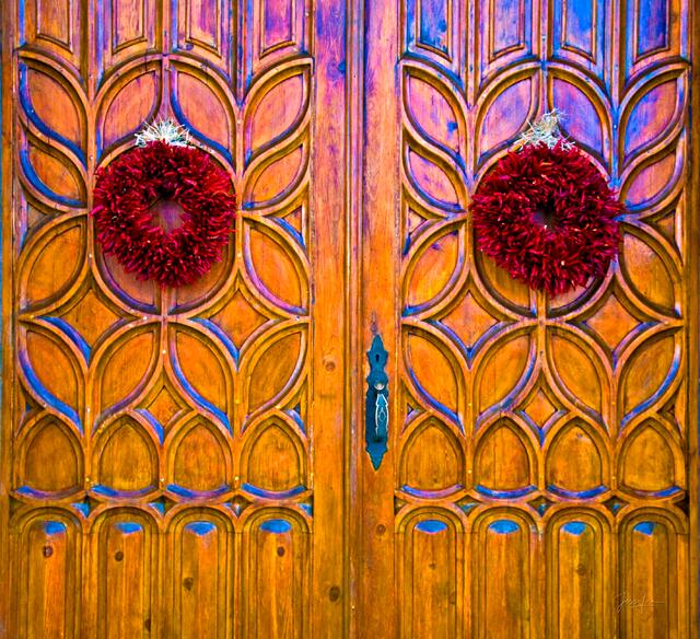 Fine art photography print of a close-up pair of ochre double doors with red chile wreaths in Santa Fe, New Mexico, featuring high-contrast colors and weathered