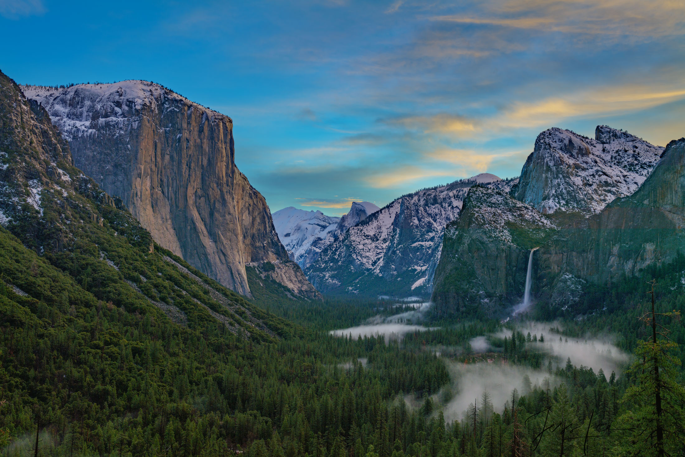 An early morning capture of the serene Yosemite Valley, adorned with patches of ethereal fog and wispy clouds against a clearing...