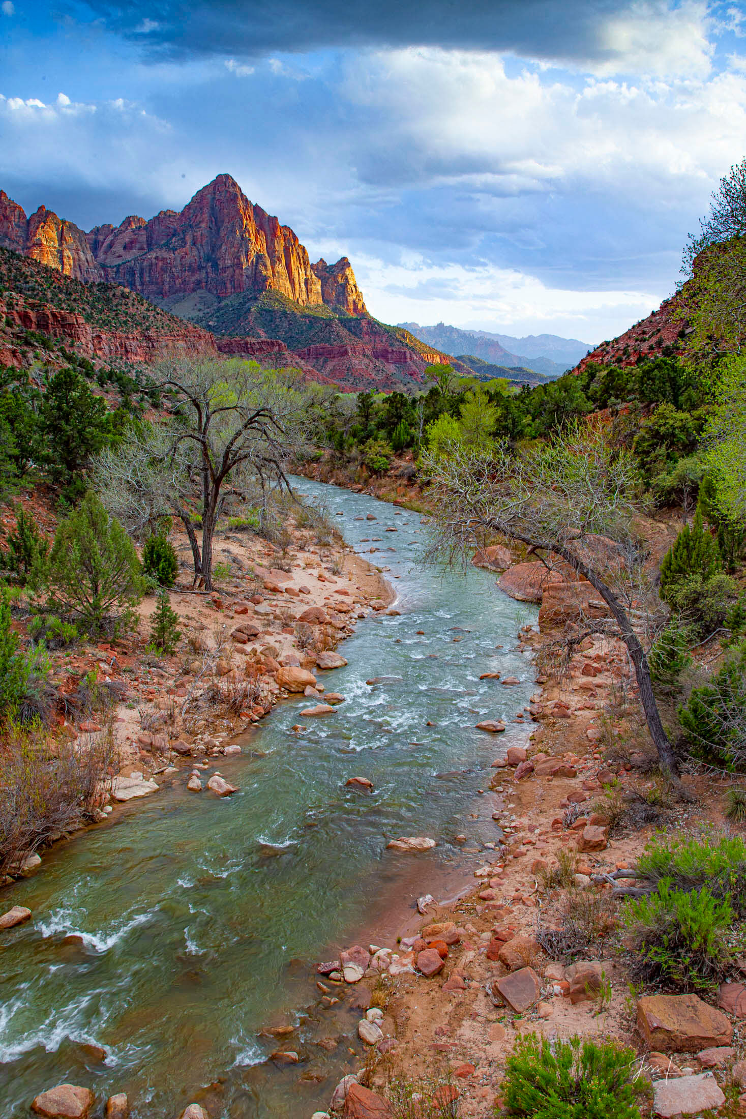 Eternal Watchman The Virgin River curves through a rocky valley toward a glowing red mountain peak under a dramatic, cloudy sky.